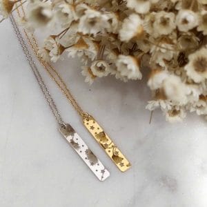 Dwa naszyjniki celebrytka prostokątne z wyciętymi gwiazdkami w formie srebrnej i pozłacanej. Naszyjniki leżą na marmurowej podstawce