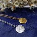 Dwa naszyjniki celebrytka z kwiatami w formie srebrnej i pozłacanej. Naszyjniki leżą na lapis lazuli