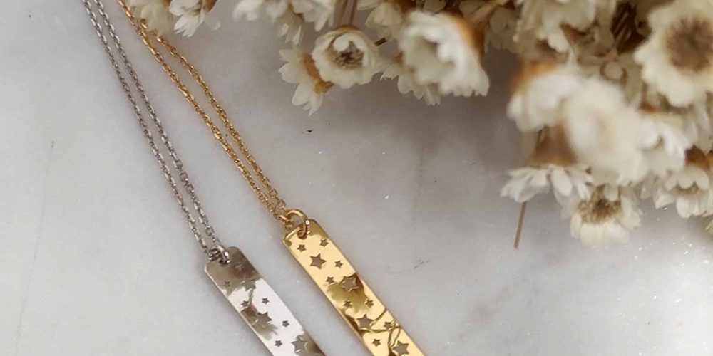 Dwa naszyjniki celebrytka prostokątne z wyciętymi gwiazdkami w formie srebrnej i pozłacanej. Naszyjniki leżą na marmurowej podstawce
